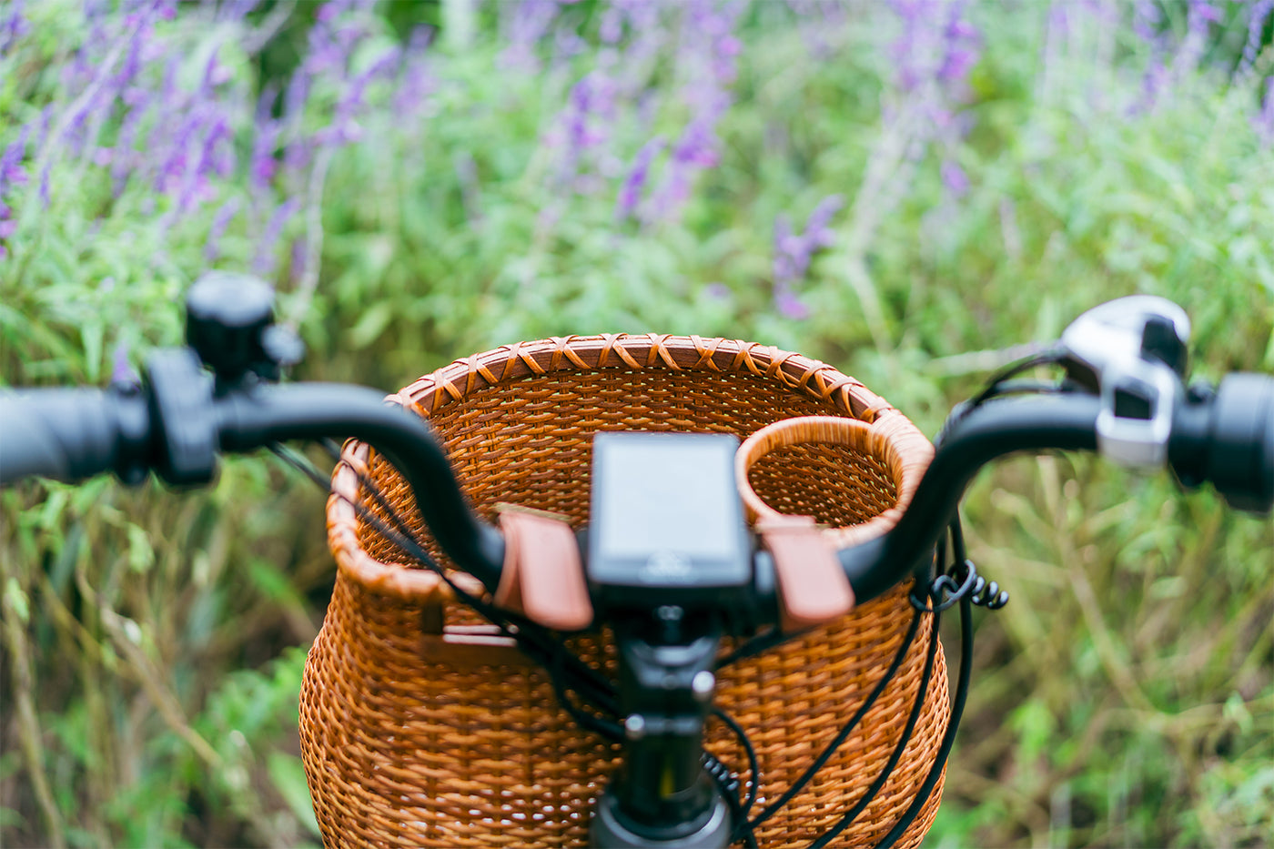 Rattan bicycle basket on handlebars with cup holder, set against purple flowers background