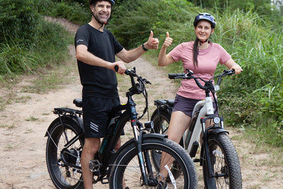 Two cyclists giving a thumbs up on electric bikes in a forested area.