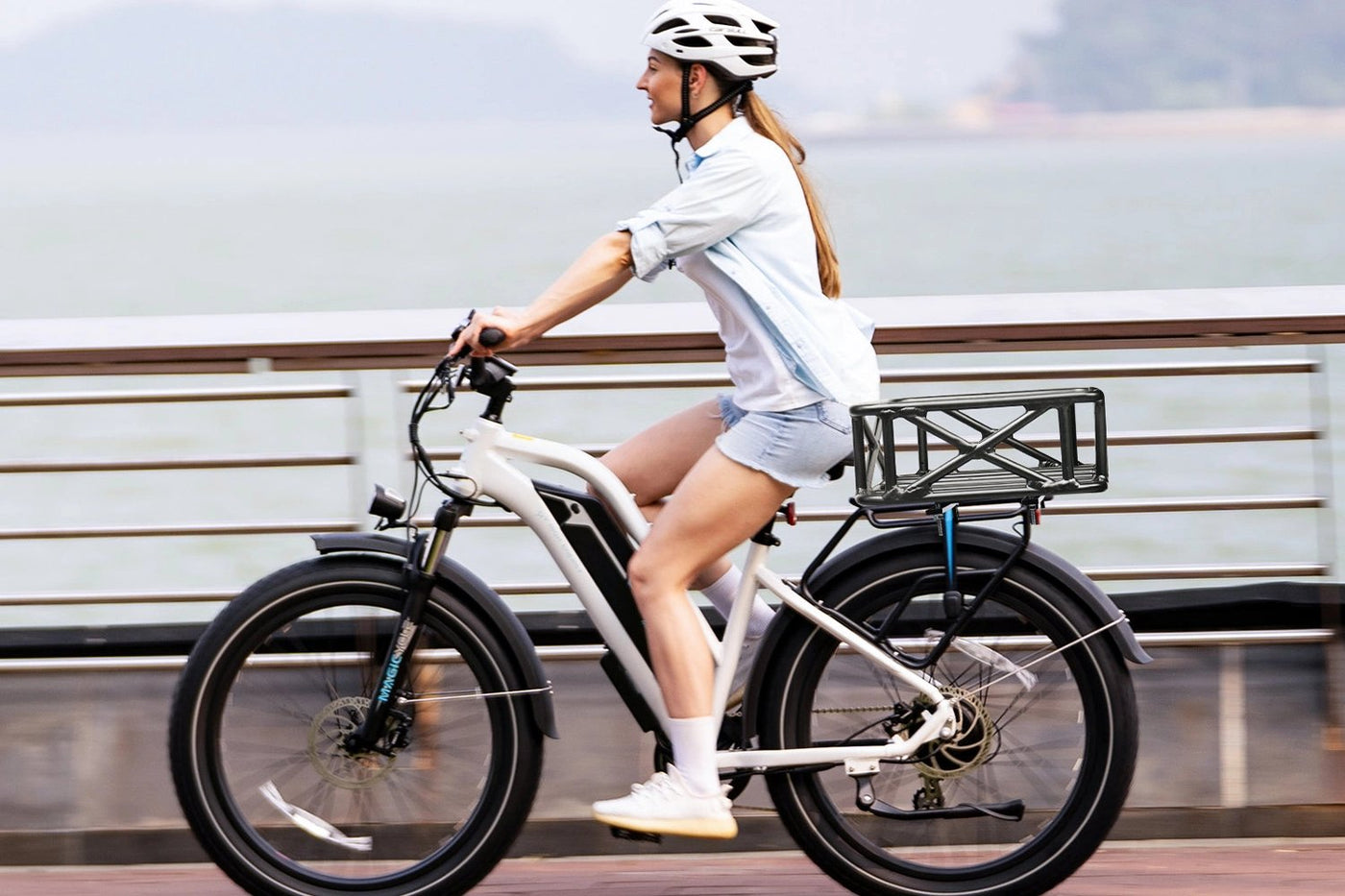 Woman riding Magicycle ebike with large black rack basket along waterfront.
