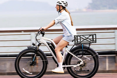 Woman riding Magicycle ebike with large black rack basket along waterfront.