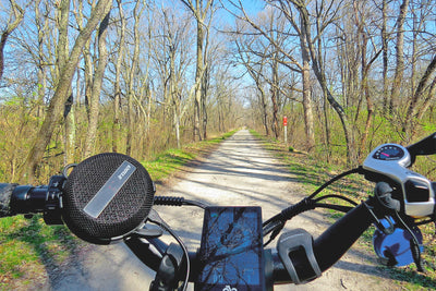 Bluetooth e-bike speaker on handlebars with scenic nature trail in background.
